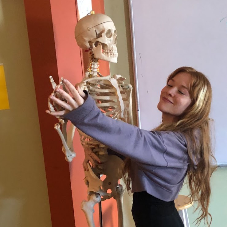 A young redheaded woman with white streaks coming from her bangs down to the tips of her long hair dances soulfully with a skeleton